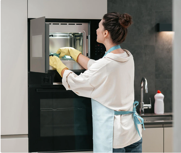 Woman cleaning an oven in a modern kitchen while wearing gloves and an apron.