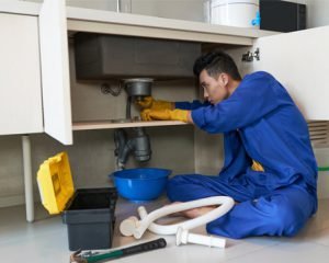 Plumber repairing a kitchen sink pipe under the counter with tools and equipment.