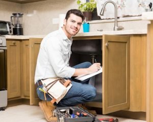 Plumber inspecting a kitchen sink and taking notes beside a toolbox.